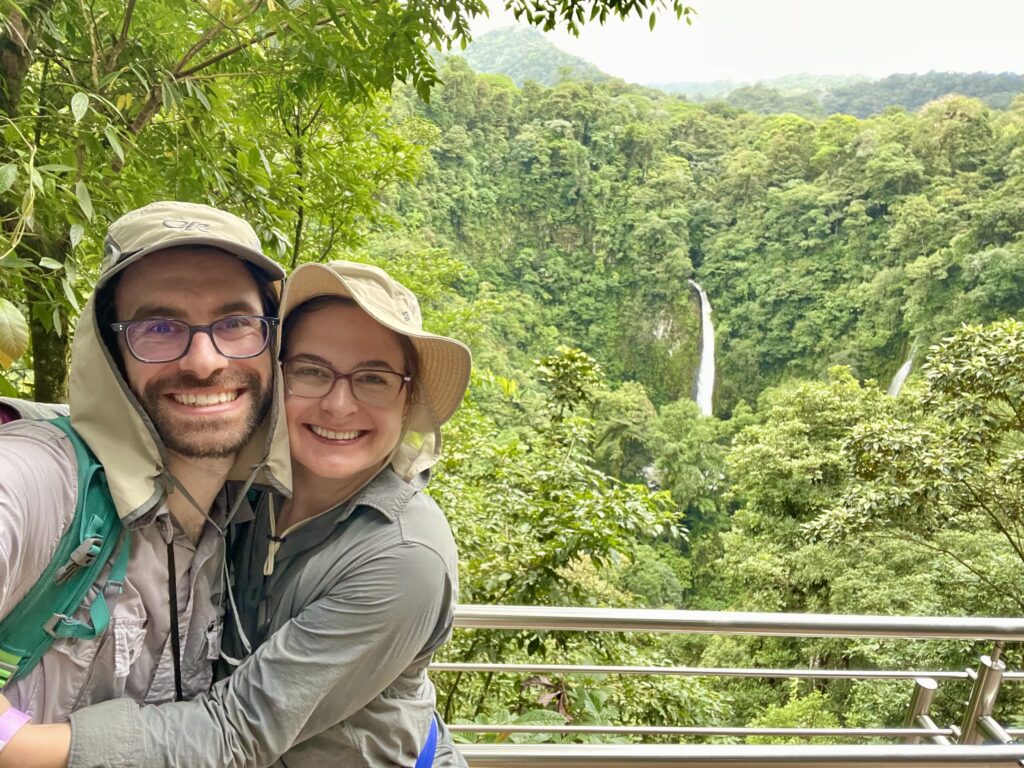 two people wearing safari hats smile in foreground and you see a waterfall in the background surrounded by jungle