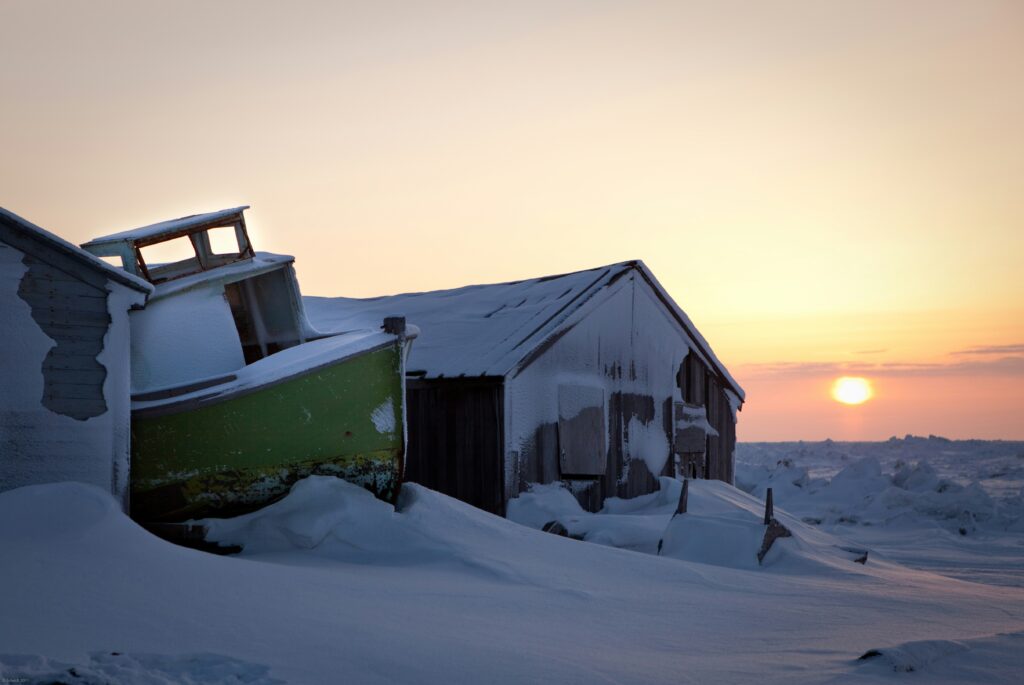 Picture of a big, green boat covered in snow and leaning up against a large shack-like house that is also covered in snow. Sun setting in the background over miles of ice and snow.
