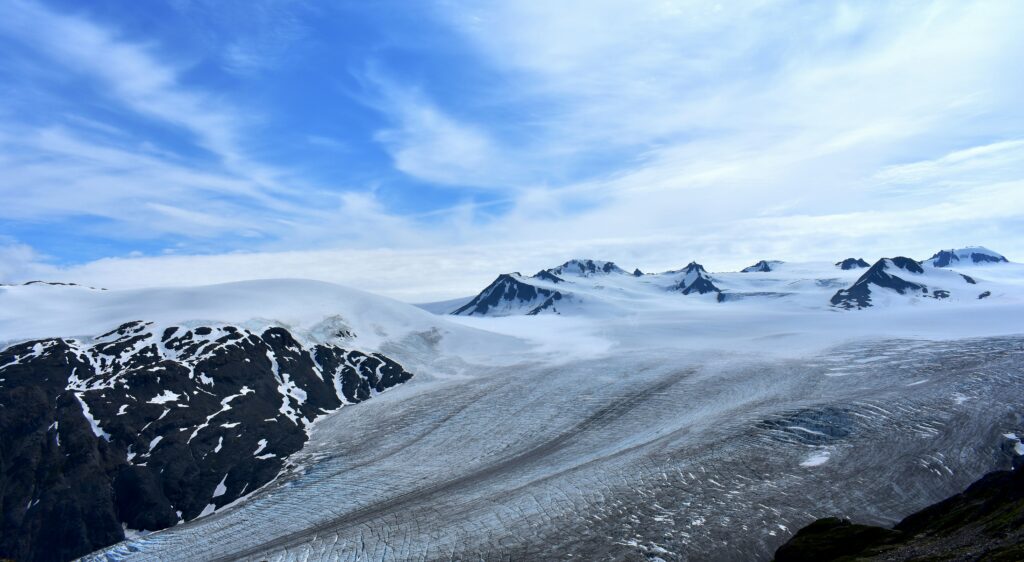 Picture of small mountain peaks and a large swatch of what looks like snow but is actually a glacier! Blue sky with wispy white clouds.