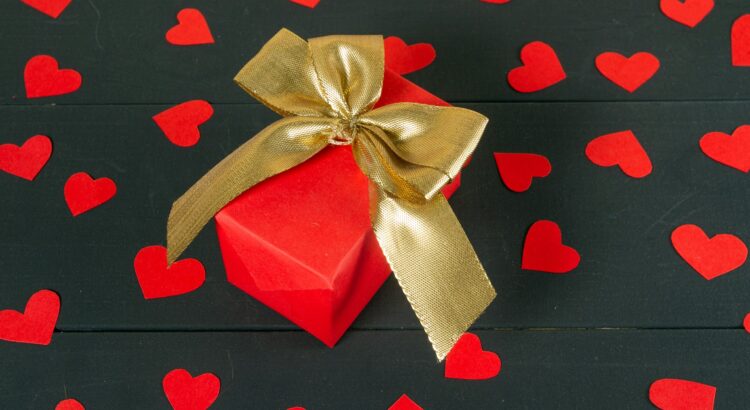 picture of red gift box with gold bow on top surrounded by red paper hearts on black paper