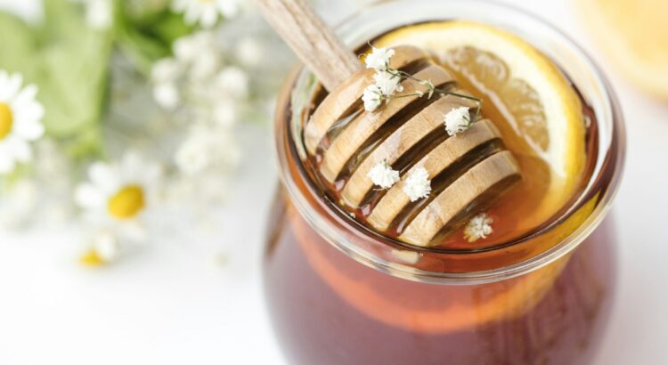 Photo of honey pot with stirrer and lemon slice in it. Out of focus, in the background are small white flowers with yellow centers and some baby's breath flowers.