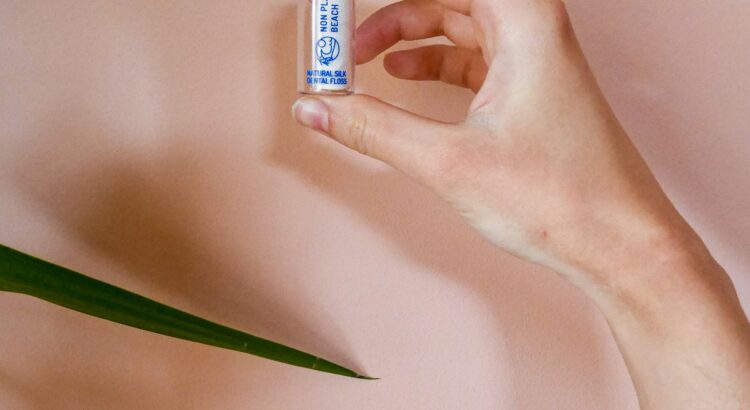Hand holding a glass sustainable floss jar in front of a peach background. There are also some long green leaves.