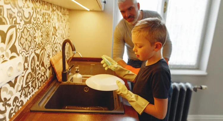 Blond haired boy washes a white dish as father watches and smiles