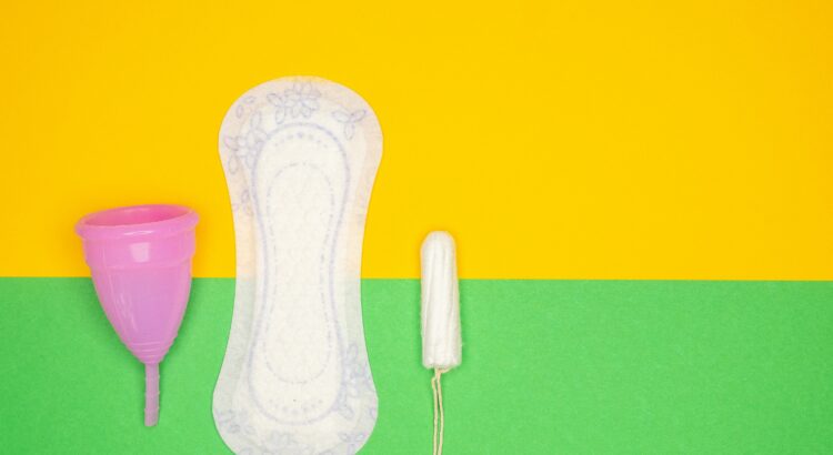 Photo of a half yellow, half green background with a menstrual cup, a pad and a tampon placed on the lower left-hand side