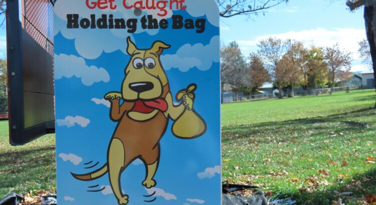 photo of a sign. on the sign is a dog holding a poop bag. the sign asks dog owners to clean up after their pets.