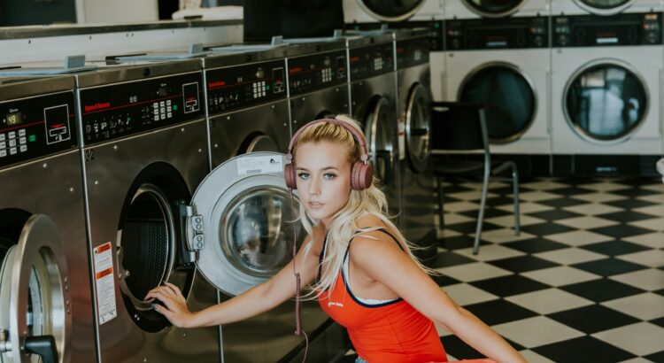 Woman in red tank top, blond hair, shorts, sneakers, holding a red laundry basket and crouching in front of a laundry machine in a laundromat