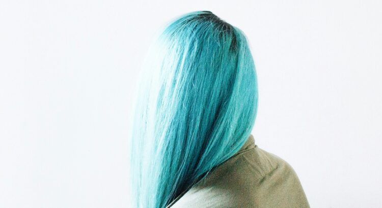 Photo of person with long, blue hair facing away from the camera and wearing an olive green shirt. White background.