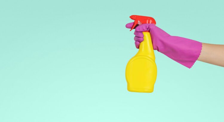 Arm covered in a pink dish washing glove, holding a yellow spray bottle in front of a light blue background